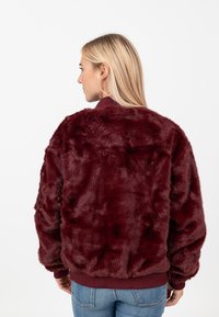 Giacca in pelliccia sintetica bordeaux con una texture morbida, vestibilità rilassata, polsini a coste e cinturino in vita, abbinata a jeans blu.