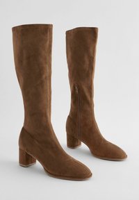 Bottes hautes en daim marron jusqu'au genou avec bout carré, talon bloc et fermeture éclair latérale. Texture douce et design épuré.