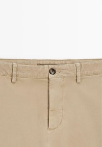 Taille de pantalon beige avec fermeture à bouton, passants de ceinture et surpiqûres visibles sur la braguette, sur fond blanc.