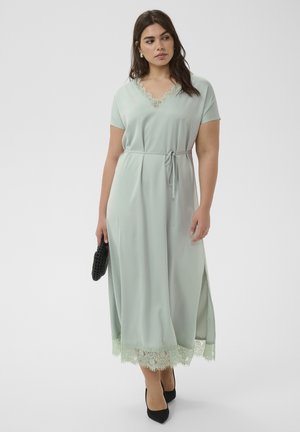 Vrouw draagt een lichtgroene v-halsjurk met kantafwerking, een ceintuur in de taille, houdt een kleine zwarte clutch vast en draagt zwarte hakken.