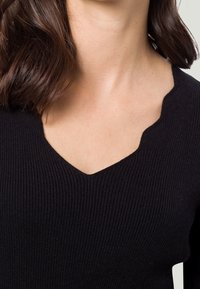 Pull noir à côtes avec un décolleté en V profond et un tissu texturé. La coupe est ajustée, mettant en valeur le décolleté et les épaules.
