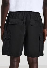 I pantaloni cargo neri presentano una vestibilità ampia, una cintura elastica, due tasche laterali e due tasche posteriori con patta. Il tessuto ha una consistenza liscia.
