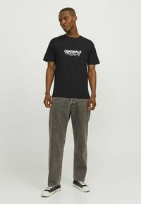 Camiseta negra con gráfico blanco "ORIGINALS" en el pecho, combinada con pantalones vaqueros grises holgados y zapatillas negras con suelas blancas.