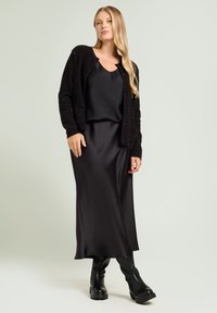 Schwarze Strickjacke aus strukturiertem Stoff mit Knopfschluss, über einem schwarzen satinierten Tanktop und einem eleganten schwarzen Maxirock getragen, kombiniert mit knielangen Stiefeln.