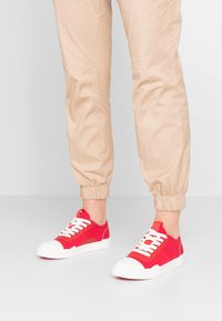 Baskets en toile rouges avec des semelles et des lacets en caoutchouc blanc, présentant une forme de bout arrondi et une finition en tissu texturé. Portées avec un pantalon beige clair.