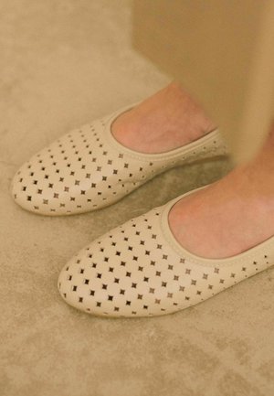Ballerine in pelle beige con motivi a stella perforati, punta arrotondata e bordi elasticizzati per una vestibilità sicura, posizionate su una superficie testurizzata.