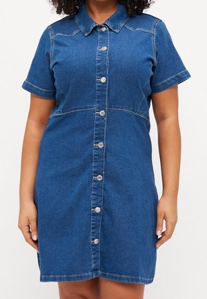 Kvinde iført en kortærmet blå denim skjortekjole med krave, stående mod en ensfarvet hvid baggrund.