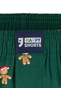 Groene shorts met een peperkoekenmannetjespatroon, elastische tailleband en een marineblauw label met "HAPPY SHORTS" in kleurrijke tekst.