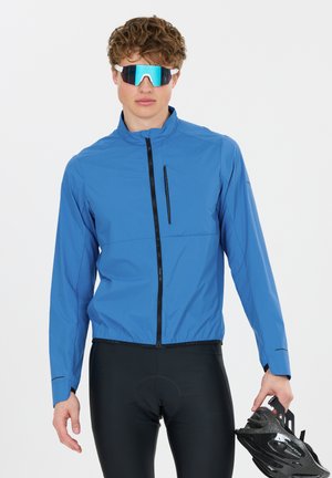 Junger Mann trägt eine blaue Fahrradjacke und reflektierende Sonnenbrille, hält einen schwarzen Fahrradhelm in einer Hand.