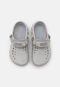 Sabots en caoutchouc gris avec design ouvert, sangle beige ajustable et trous de ventilation. Semelle intérieure texturée pour le confort. Bords nervurés.