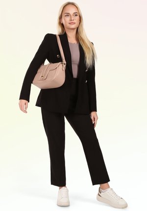 Blonde Frau trägt schwarzen Anzug, mauvefarbenes Top, weiße Sneakers, trägt eine beige Umhängetasche, steht vor hellem Hintergrund.