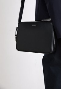 Czarna, skórzana, prostokątna torebka na ramię z regulowanym paskiem, gładką fakturą, wytłoczonym logo i detalem zapinania na suwak.