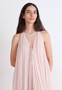 Lys rosa halterneck-kjole laget av flytende stoff, med en samlet neckline og knyttet foran. Akkompagnert med et enkelt, rundt halskjede.