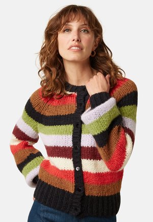 Mujer con cabello rizado y castaño que lleva un cárdigan de punto a rayas multicolor con botones, posando con una mano cerca del cuello.