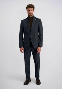 Man in een donkergrijs pak met een bruine coltrui en donkerbruine nette schoenen, staand tegen een effen lichte achtergrond.