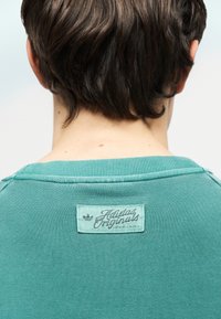 Κοντά σκούρα καστανά μαλλιά σε άτομο που φοράει μπλούζα Adidas Originals σε τιρκουάζ χρώμα, με θέα από πίσω, εστιάζοντας στην ετικέτα στον λαιμό.