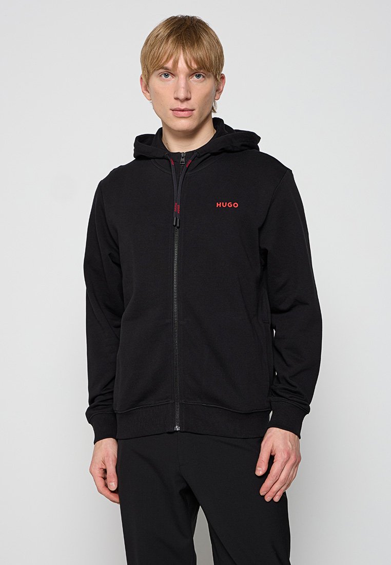 Haut à zip noir en coton doux, avec une fermeture éclair à l'avant et une capuche. Logo "HUGO" en rouge sur la poitrine à gauche. Texture lisse.