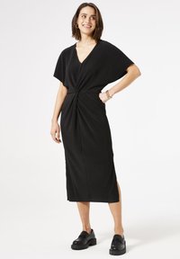 Vestido negro de punto con mangas cortas, escote en V y detalle frontal retorcido. Presenta aberturas laterales y una longitud midi recta. Se lleva con zapatos de plataforma negros.