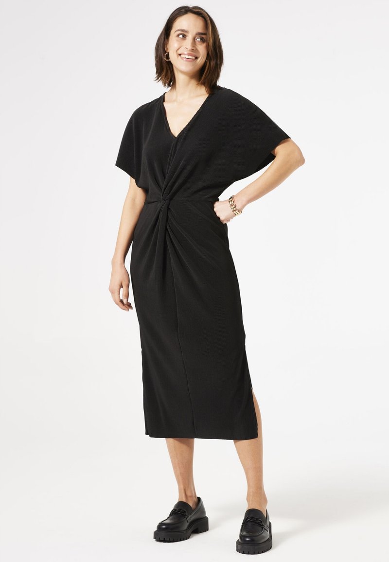 Vestido negro de punto con mangas cortas, escote en V y detalle frontal retorcido. Presenta aberturas laterales y una longitud midi recta. Se lleva con zapatos de plataforma negros.