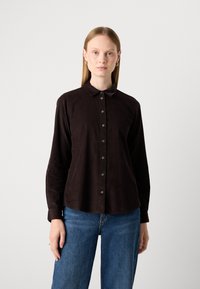 Woman with straight blonde hair wearing gold hoop earrings, a black buttoned corduroy shirt, and blue jeans, standing against a plain background.
