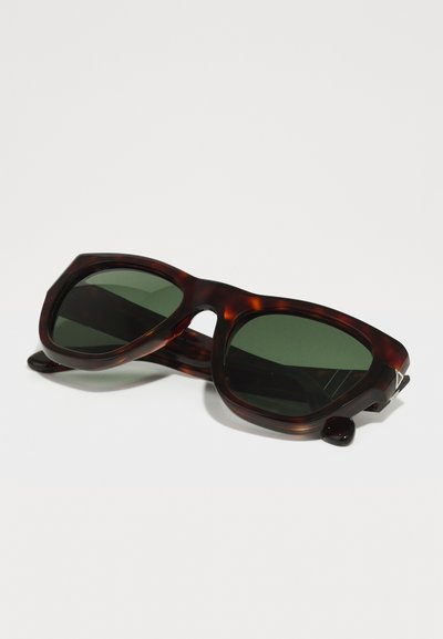 Óculos de sol de padrão tartaruga com lentes verdes, formato angular, armação de plástico texturizado e detalhes em metal junto às dobradiças.