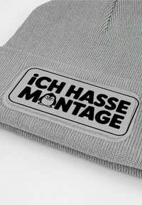 Gray knit beanie featuring a rectangular patch with a cartoon penguin and the text "iCH HASSE MONTAGE" in bold black letters.