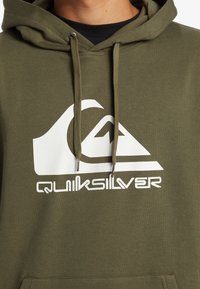 Khaki hoodie with a front kangaroo pocket, drawstrings, and a white Quiksilver logo on the chest. Cotton material, smooth texture.