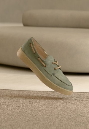 Grüner Bootsschuh aus Wildleder mit beiger Schnürung und seitlichen Aussparungen, ausgestattet mit einer strukturierten Gummisohle und schlankem Design, schwebend über einer strukturierten Oberfläche.