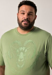 Camiseta verde de manga corta de algodón con un estampado texturizado de la cabeza de un carnero y un patrón decorativo de follaje. Cuello redondo, corte relajado.