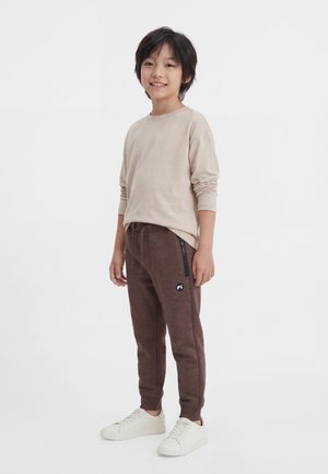 Garçon souriant debout, portant une chemise beige à manches longues, un pantalon de jogging marron avec des poches zippées, et des baskets blanches sur un fond uni.