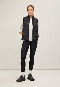 Schwarzte gepolsterte Weste mit hohem Kragen, Reißverschluss und seitlichen Taschen. Getragen über einem weißen Langarmoberteil und kombiniert mit schwarzen Leggings und Turnschuhen.