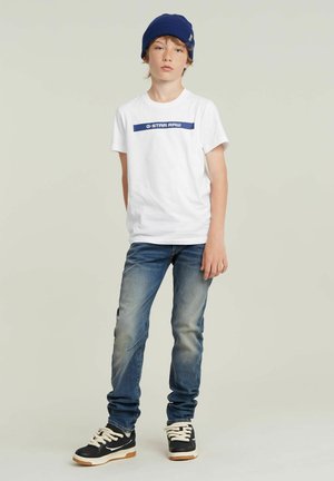 Jongen in een blauwe beanie, wit G-Star Raw t-shirt, vervaagde blauwe spijkerbroek en zwarte sneakers, staand tegen een effen lichtgrijze achtergrond.