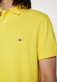 Keltainen polo-paita, jossa on tekstuuri, kolme nappia ja pieni, punavalkosininen kirjailtu logo vasemmassa rinnassa.