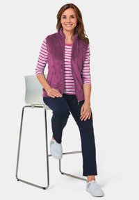Fuchsia Fleece-Weste über einem rosa-weiß gestreiften Hemd, kombiniert mit marineblauen Hosen und weißen Sneakern, sitzt auf einem Metallhocker.