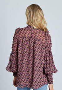 Blusa morada con estampado, con mangas abullonadas, que presenta detalles de volantes y un corte holgado. Tela ligera y semitransparente.