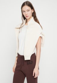 Νεαρή γυναίκα με μακριά καστανά μαλλιά, που φοράει λευκό μπλουζάκι, κρεμ φούτερ περασμένο στους ώμους και καφέ παντελόνι φόρμας GAP.