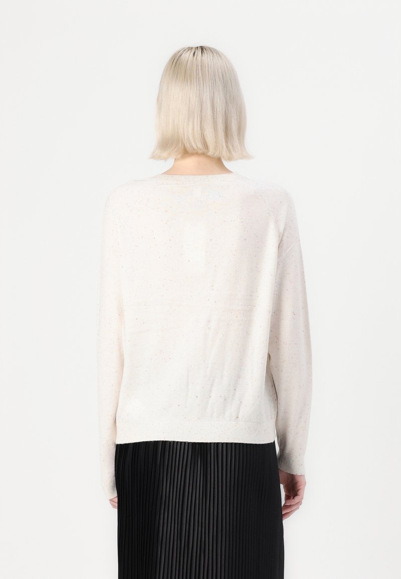 Hellbeiger Pullover mit gesprenkelter Textur, langen Ärmeln und rundem Ausschnitt. Getragen über einem schwarzen, plissierten Rock, zeigt die Rückansicht.