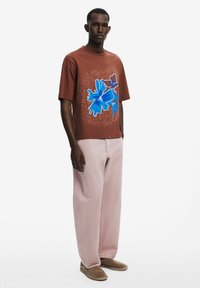 T-shirt marron à manches courtes avec un design graphique abstrait bleu, associé à un pantalon ample rose clair et des chaussures beiges à enfiler.