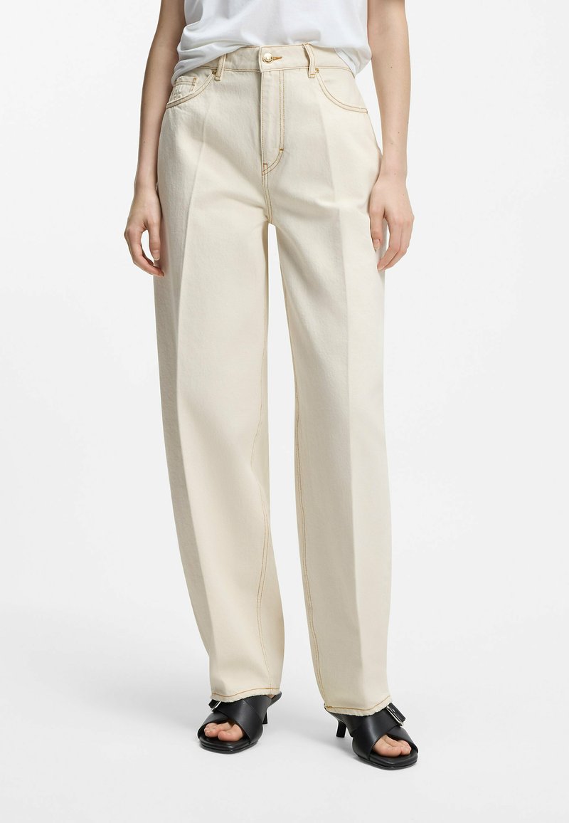 Kvinde iført off-white brede bukser, hvid indstukket skjorte og sorte hælede sandaler mod en ensfarvet baggrund.