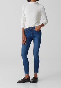 Jeans blu scuro aderenti con una leggera elasticità, abbinati a un top bianco a maniche lunghe con texture. Mocassini marroni completano l'outfit.