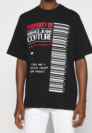 T-shirt noir Versace Jeans Couture avec texte rouge et blanc et un grand graphique de code-barres vertical sur le devant, porté avec un pantalon blanc.