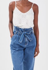 Camisole en dentelle blanche avec un décolleté en V, associée à un pantalon en denim bleu taille haute avec une ceinture à nouer et des poches latérales.