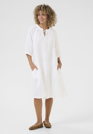 Robe en lin blanc avec un col à volants, des manches trois-quarts et des poches. Présente une patte de boutonnage centrale avec des détails en dentelle.