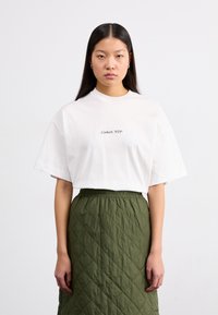 Bílý bavlněný trikotaž s krátkými rukávy, s černým textem "Carhartt WIP". V kombinaci s prošívanou sukni olivové zelené barvy s elastickým pasem.
