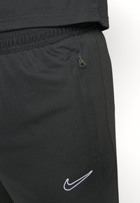 Sorte atletiske bukser i et tekstureret stof, med en elastisk talje og en lynlåslomme, der har et hvidt Nike-logo på venstre ben.