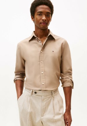 Junger Mann mit hochgekrempelten Ärmeln, der ein beiges Hemd mit Knöpfen und hellbeige plissierte Hosen trägt, steht mit den Händen in den Taschen.