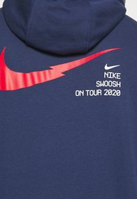 Navy hoodie featuring a red Nike Swoosh graphic and white text "NIKE SWOOSH ON TOUR 2020." Soft fabric with a kangaroo pocket design.