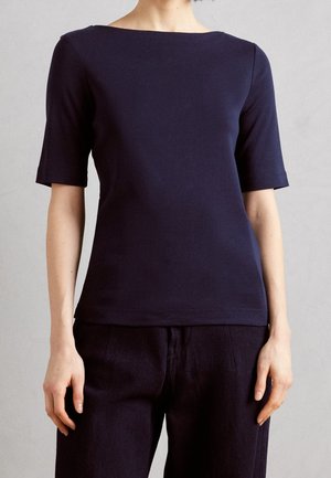 Kobieta w dopasowanym, granatowym topie z krótkim rękawem i dekoltem w łódkę oraz w dopasowanych ciemnych spodniach na neutralnym tle.