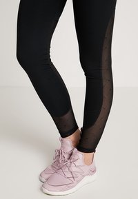 Svarta leggings med genomskinliga nätpaneler, med en texturerad design. Kombineras med ljusrosa träningsskor med en texturerad ovandel.