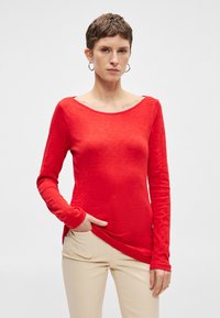 Hosszú ujjú piros felső, textúrált anyagból, széles nyakkivágással és diszkrét oldalsliccekkel, világos bézs nadrággal párosítva.